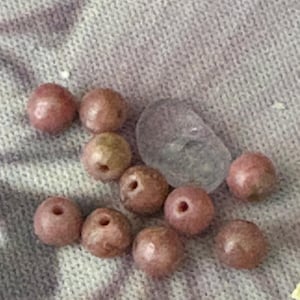 May include: A collection of pinkish-brown beads with holes, alongside a translucent, irregularly shaped stone. The beads appear to be made of a natural material. The items are arranged on a textured, light purple surface.