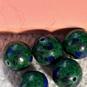 Puede incluir: Cinco cuentas redondas con una mezcla de colores verdes y azules. Cada cuenta tiene un agujero perforado. Las cuentas tienen un acabado pulido y brillante. Las cuentas son probablemente para la fabricación de joyas o manualidades.