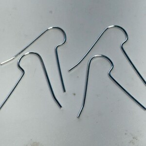 Può includere: Quattro orecchini minimalisti color argento. Ogni orecchino presenta un design a gancio curvo con un'estremità dritta e appuntita. Gli orecchini sono esposti su una superficie di colore chiaro.