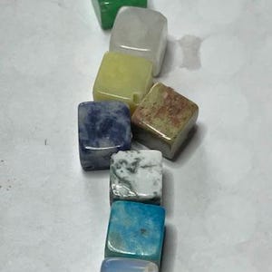 Puede incluir: Una colección de siete gemas cuadradas y coloridas. Las gemas están dispuestas verticalmente e incluyen variaciones verdes, claras, amarillas, azules y blancas. Las piedras tienen un aspecto pulido y varían en color y patrón.