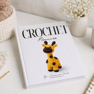 Puede incluir: Un libro de tapa dura blanco titulado Crochet Planner con un gráfico de una jirafa tejida a ganchillo amarilla y marrón en la portada. La portada presenta el texto Crochet Planner y By: Seluanna Studio. El libro está rodeado de hilo y ganchillos.