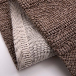 May include: A brown rug with a thick, textured, woven surface and a folded corner revealing a light beige, tightly woven fabric backing. The edge of the rug is finished with a light beige herringbone pattern trim along the folded seam.