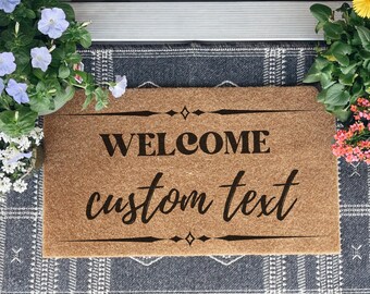 May include: A rectangular coir doormat with the words "WELCOME custom text" in black script. The mat is bordered by decorative lines and diamonds. The mat is on a patterned rug, with potted flowers on either side.