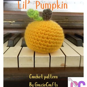 Peut inclure: Une citrouille orange crochetée avec des feuilles vertes et une tige marron posée sur un clavier de piano. Le texte "Lil' Pumpkin" est au-dessus et "Crochet pattern By GracieCrafts" est en dessous.
