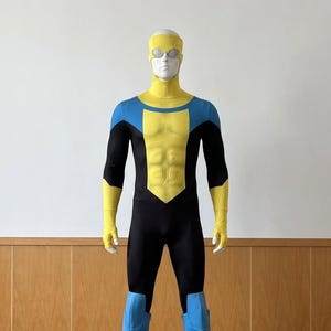 Puede incluir: Disfraz de superhéroe negro, amarillo y azul sobre un maniquí. El traje presenta un torso amarillo, hombros azules, botas azules y guantes amarillos. El maniquí lleva una capucha amarilla y gafas.