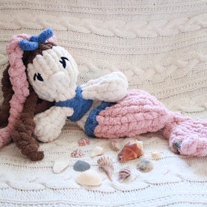 May include: A handmade plush mermaid doll with a white head, blue top, and pink tail. The doll has brown and pink hair and is lying on a white, textured blanket. Seashells are scattered around the doll.