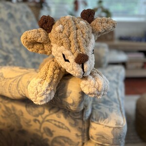 May include: A handmade plush deer toy with a textured, fluffy appearance. The deer features a light brown and cream color scheme, with dark brown antlers and a snout. The toy is resting on a patterned armchair, showcasing its soft, cuddly design.