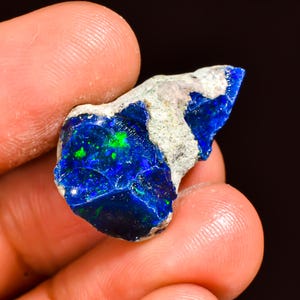 May include: A vibrant, raw opal gemstone with a striking display of deep blue and green colors. The opal is held in a hand, showcasing its natural, rough texture and unique color patterns. The stone has a mix of blue, green, and white hues.
