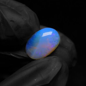Könnte beinhalten: Ein ovaler Cabochon-Opal-Edelstein mit einem durchscheinenden Körper und lebhaften Blitzen in Blau, Orange und Violett. Der Stein hat eine glatte, polierte Oberfläche, die das Licht reflektiert und ein Farbspiel über seine abgerundete Form vor einem dunklen Hintergrund zeigt.