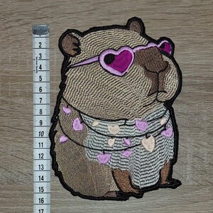 May include: Embroidered patch of a brown rodent wearing heart-shaped sunglasses and a sweater decorated with heart-shaped garlands. The patch is approximately 15 cm in height.