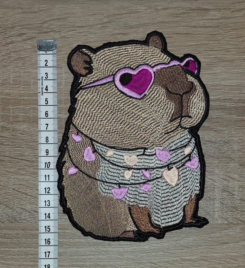 May include: Embroidered patch of a brown rodent wearing heart-shaped sunglasses and a sweater decorated with heart-shaped garlands. The patch is approximately 15 cm in height.