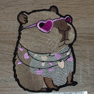 May include: Embroidered patch of a capybara wearing heart-shaped sunglasses and a garland of hearts. The capybara is in shades of brown, tan, and purple, with a black outline. A ruler is placed below the patch.