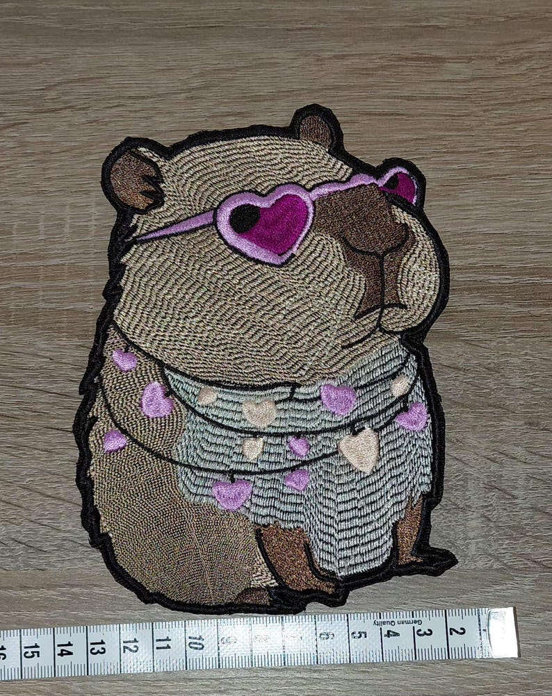 May include: Embroidered patch of a capybara wearing heart-shaped sunglasses and a garland of hearts. The capybara is in shades of brown, tan, and purple, with a black outline. A ruler is placed below the patch.
