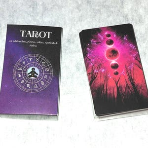 May include: A tarot card deck with a purple and black box featuring the word "TAROT" and astrological symbols. The deck of cards has a vibrant image of a red and pink celestial scene with trees.
