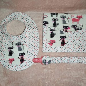 Pode incluir: Um conjunto para bebé composto por um babete, um clipe para chupeta e uma capa para muda-fraldas. Os artigos são brancos com um padrão de gatos rosa, cinzentos e pretos e estrelas rosa e cinzentas. O babete tem um desenho circular de gato.
