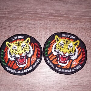Op de afbeelding: Twee ronde patches met een tijgerontwerp. De tijger is geel en wit met rode accenten, tegen een zwarte achtergrond met oranje strepen. Tekst bevat "NTM 2025" en "PORTUGAL .BEJAAIRBASE.301SQN".