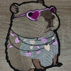 Op de afbeelding: Geborduurde patch met een capibara met een hartvormige zonnebril en een slinger van harten. De patch heeft een gedetailleerd ontwerp met bruine, roze en zwarte kleuren. De capibara is de centrale focus.