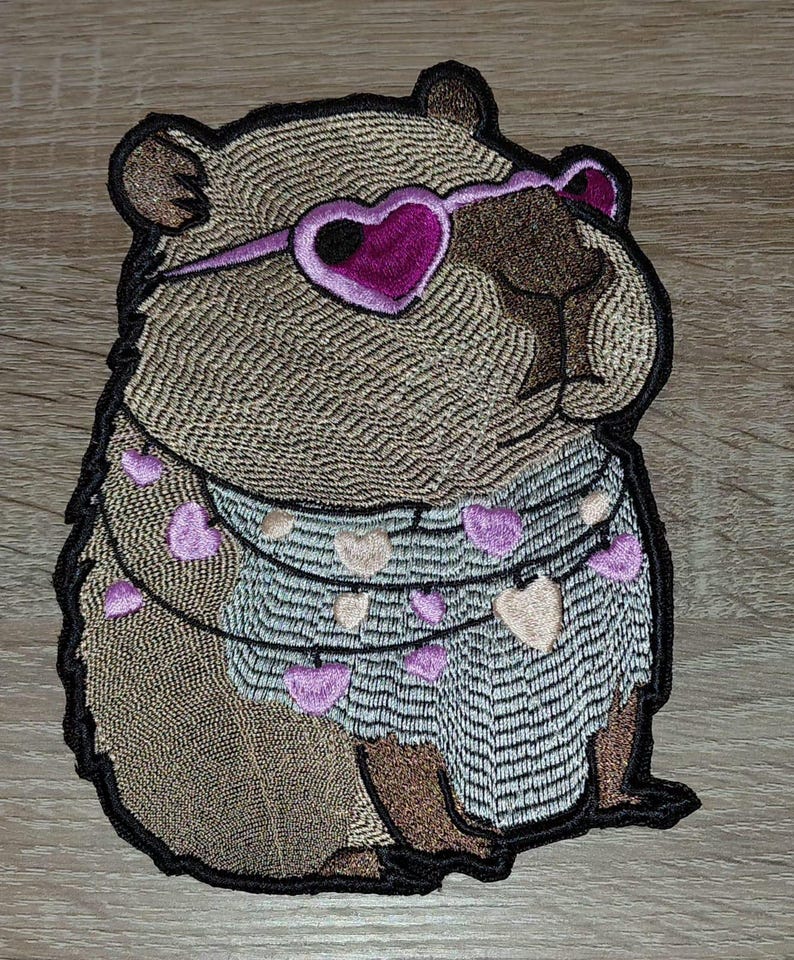 May include: Embroidered patch of a capybara wearing heart-shaped sunglasses and a garland of hearts. The patch has a detailed design with brown, pink, and black colours. The capybara is the central focus of the image.