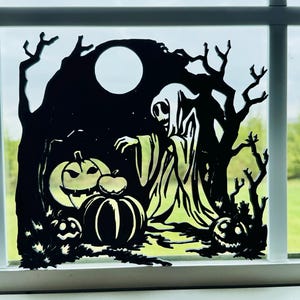 May include: A black silhouette window cling featuring a hooded figure standing in a forest with a full moon above. The scene includes three carved jack-o'-lanterns with glowing eyes and mouths positioned near the base of the trees.