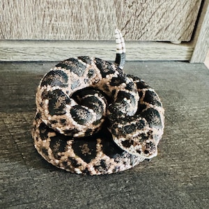May include: A coiled rattlesnake figurine featuring a textured body with a pattern of dark brown and black diamond-shaped blotches on a tan background. The snake has a distinct triangular head and a raised, segmented rattle at the tail tip.