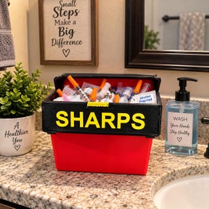 May include: A red and black sharps container with the word SHARPS printed in large yellow letters on the front. The container is filled with various medical syringes, needles, and an alcohol prep pad, featuring a bright yellow latch mechanism.