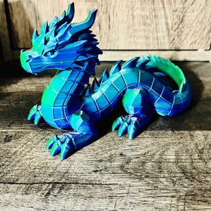 May include: A 3D printed articulated dragon figurine featuring a vibrant gradient of blue, green, and purple colors. The creature has a segmented body, sharp dorsal spikes, and detailed facial features with a prominent snout and pointed ears.