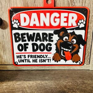 May include: A rectangular red, white, and black sign with a hanging loop at the top. It features the text DANGER, BEWARE OF DOG, and HE'S FRIENDLY... UNTIL HE ISN'T! A cartoon illustration of a black and brown dog with a red tongue is included.