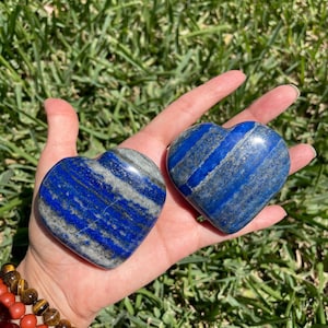 May include: Two heart-shaped lapis lazuli stones with blue and gray veining are held in a hand. The stones are polished and smooth, with a natural, organic appearance. The background is blurred green grass.