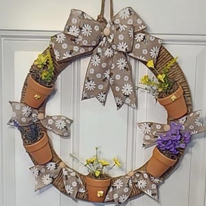May include: A decorative wreath featuring a circular woven base adorned with small terracotta pots holding colorful faux flowers. The wreath is embellished with a large bow and smaller bows made of fabric printed with white daisies and bees.
