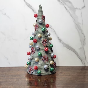 May include: A conical Christmas tree decoration crafted from coiled sage green rope. The surface is adorned with small, shiny spherical ornaments in red, green, silver, and cream, interspersed with miniature plaid fabric bows throughout.