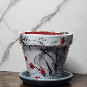 May include: A white plant pot with a black and white botanical print featuring dragonflies and red poppy flowers. The pot has a wide rim, a red interior, and sits on a matching dark gray circular saucer with a raised edge and a tapered base.