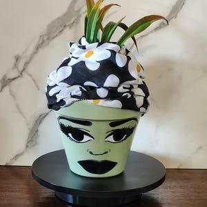 May include: A light green terracotta plant pot painted with a stylized face featuring large black eyes, long eyelashes, and dark lips. The pot is topped with a black fabric headwrap featuring a white and yellow daisy floral pattern.