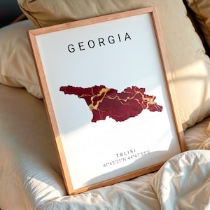 May include: A framed print featuring the country of Georgia. The map is a deep red with gold accents, set against a white background. The word "GEORGIA" is printed in black at the top, with "Tbilisi" and coordinates at the bottom.