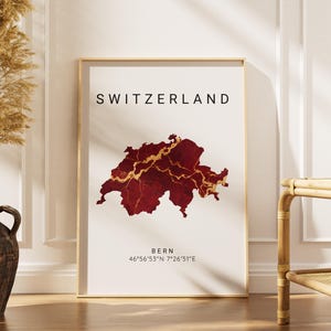 May include: A framed print featuring a map of Switzerland in shades of red and gold. The word "SWITZERLAND" is printed at the top, with "BERN" and coordinates below. The print is displayed in a room with a vase of dried plants.