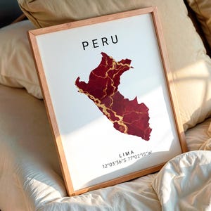 May include: A framed print featuring the map of Peru in a deep red color with gold accents. The word "PERU" is at the top, and "LIMA" with coordinates are at the bottom. The frame is a light wood color.