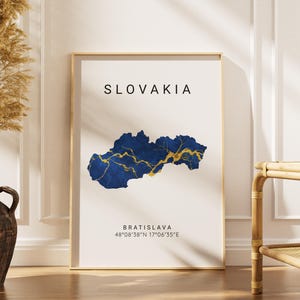 May include: A framed print featuring a map of Slovakia in shades of blue and gold. The word "SLOVAKIA" is at the top, with "BRATISLAVA" and coordinates below. The print is displayed near a vase with dried plants.