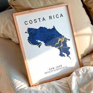 May include: A framed print showing a map of Costa Rica in blue with gold detailing. The text "COSTA RICA" is at the top, and "SAN JOSÉ" with coordinates is at the bottom. The frame is a light brown colour.