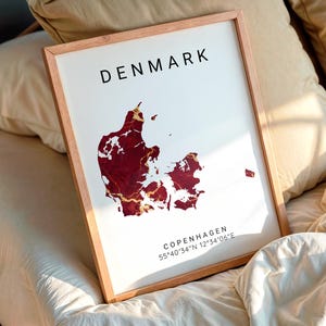 Op de afbeelding: Een ingelijste print met een kaart van Denemarken met een bordeauxrode en goudkleurige marmeren design. Het woord "DENMARK" staat in het zwart bovenaan, met "COPENHAGEN" en coördinaten eronder. De lijst is lichtbruin.