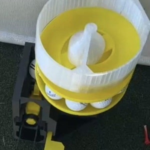 Peut inclure: Un distributeur de balles de golf doté d'un plateau circulaire jaune avec un rebord blanc translucide et une poignée centrale blanche. La base est en matériau noir avec un mécanisme à levier jaune, conçu pour contenir et libérer les balles.