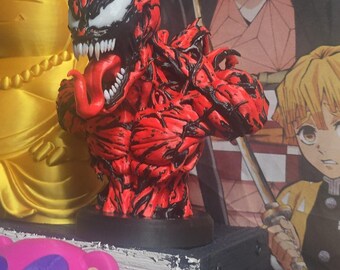 Può includere: Un busto di Carnage rosso e nero con un torso muscoloso e strutturato, una bocca spalancata con denti bianchi affilati e una lunga lingua rossa sporgente. Il personaggio ha occhi bianchi a mandorla e poggia su una base circolare nera.