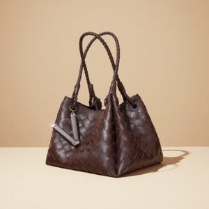 May include: A dark brown leather tote bag with a woven design. The bag has braided handles and a silver-toned charm. The bag is a trapezoid shape and has a soft, unstructured appearance.