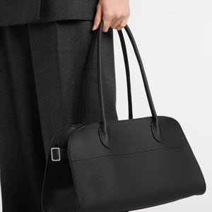 Può includere: Borsa a mano in pelle nera di forma rettangolare con due lunghi manici. La borsa ha una superficie strutturata e una fibbia color argento sul lato. La borsa è tenuta da una persona che indossa un abito grigio scuro.