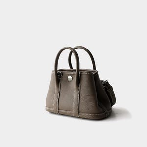 May include: A taupe-colored handbag with two rounded top handles and a detachable shoulder strap. The bag features white stitching and a silver-toned clasp. The leather bag has a structured design.