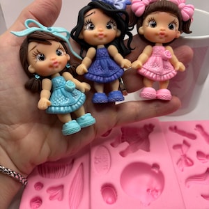 May include: Three small, handcrafted doll figurines with detailed features. Each doll has a different hairstyle and dress color: blue, purple, and pink. The dolls are held above a pink silicone mold with various shapes.