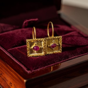 以下が含まれることがあります： 正方形のデザインの金色のイヤリングのペアで、それぞれ中央に楕円形の赤い宝石が付いています。 イヤリングは装飾的なフレームにセットされ、濃い赤色のベルベットの裏地が付いたジュエリーボックスの中に展示されています。