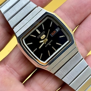 May include: A silver Seiko automatic watch with a black face and gold detailing. The watch has a rectangular face with gold hands and markers, and a date display showing "SUN 24". The watch strap is made of silver-toned metal.