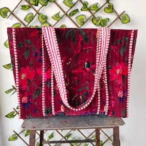 Può includere: Una borsa tote bordeaux con un motivo floreale con fiori rossi e foglie verdi. La borsa ha manici e finiture a righe bianche e rosse. La borsa è su un supporto in metallo.