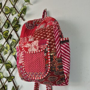 May include: A quilted backpack featuring a patchwork design in various shades of red, pink, and white. The bag includes a front pocket with a flap, side pockets, and adjustable shoulder straps, all constructed from stitched fabric panels.