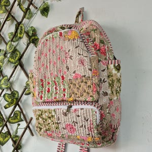 Puede incluir: Una mochila de tela acolchada con un diseño de patchwork y estampados florales en tonos rosa, amarillo, verde y beige. La mochila incluye un compartimento principal grande, un bolsillo frontal con cremallera y dos bolsillos laterales con cremalleras blancas.
