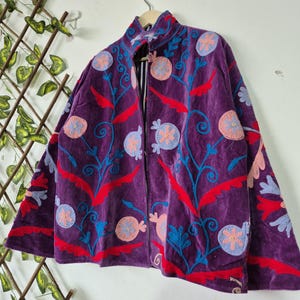 May include: A purple velvet jacket featuring vibrant embroidery in red, light blue, and peach. The design includes circular floral motifs and swirling vine patterns across the front and sleeves. The jacket has a high collar and a front opening.